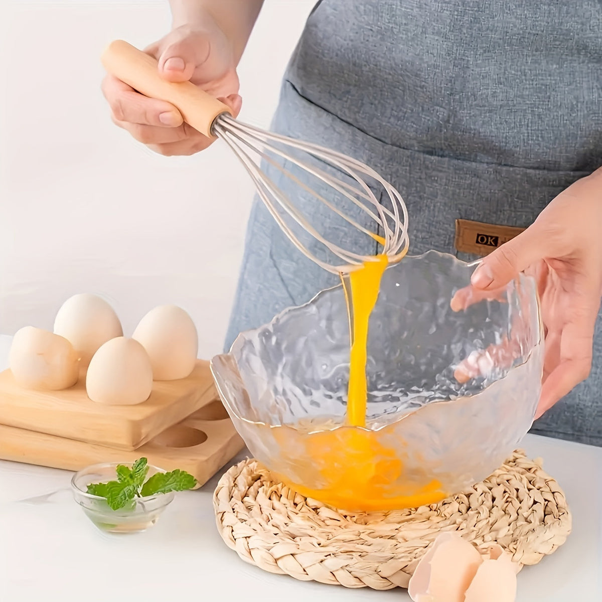 Silicone whisk with light wood handle mixing batter over a bowl on a woven mat.