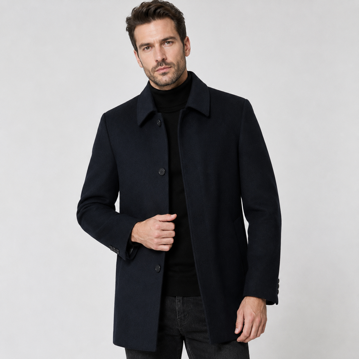 A man wearing a dark navy blue wool-blend coat over a black turtleneck sweater and dark jeans, standing against a light gray studio background.