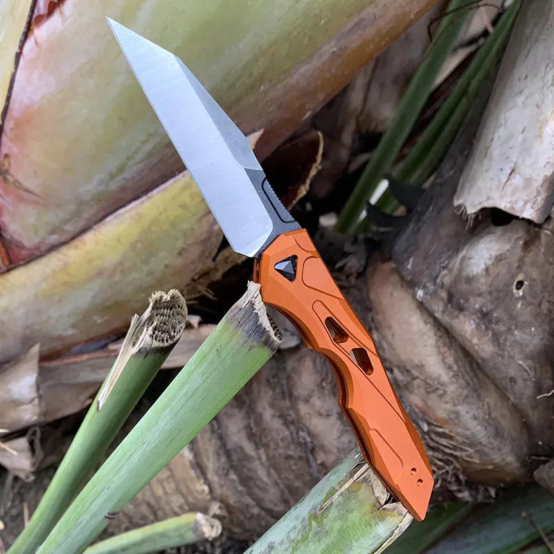 Orange auto-fold utility knife with wharncliffe blade, open among yard plants