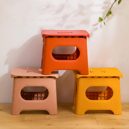 Three colorful folding stools stacked on a wooden floor with a white wall background.