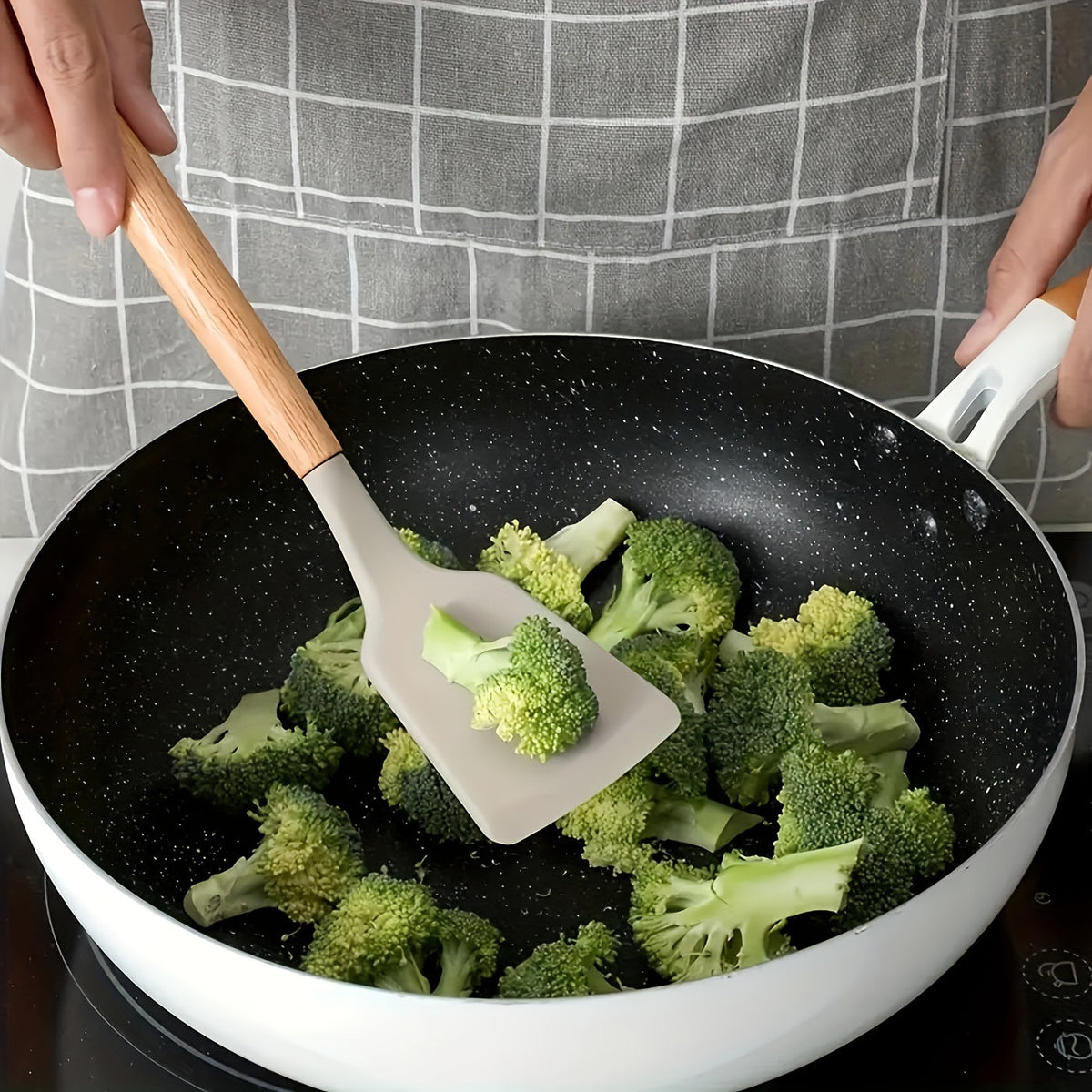 Silicone spatula with light wood handle in a nonstick skillet, holding broccoli during cooking.