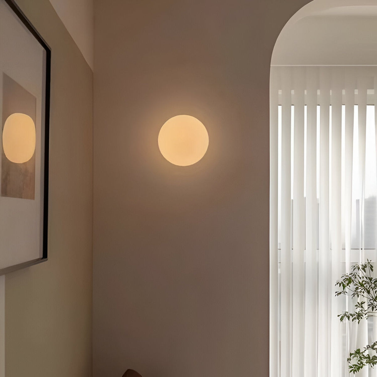Round wall light fixture on a neutral-colored wall with a framed picture and window blinds in the background.